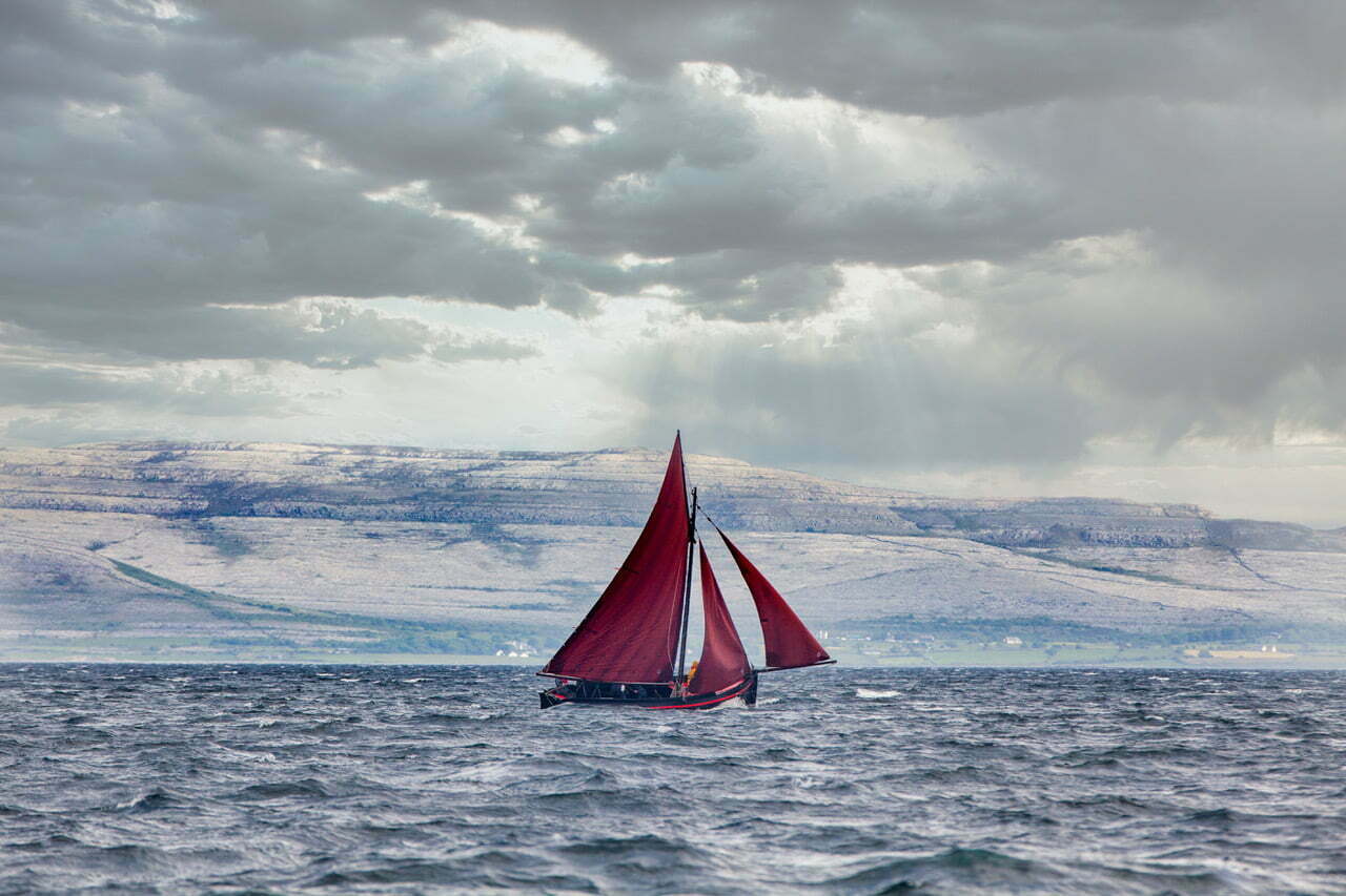 a Galway Hooker boat sailing in the blue waters of Galway Bay on a cloudy day during a Galway Girl Cruises tour.