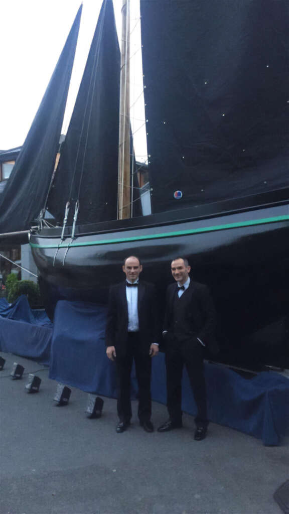 Tommy and Patrick in smart suite standing by a boat with blue sail